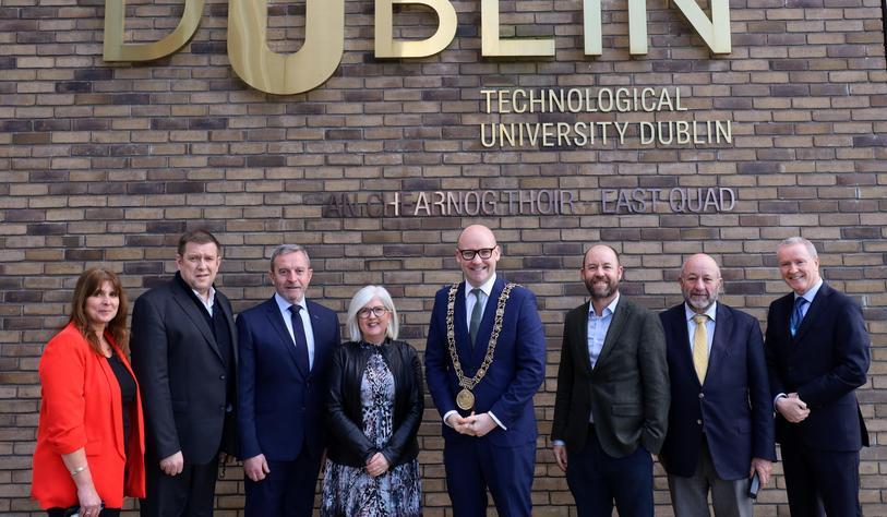 A group of people pose for a photo in front of a bronze tu dublin sign